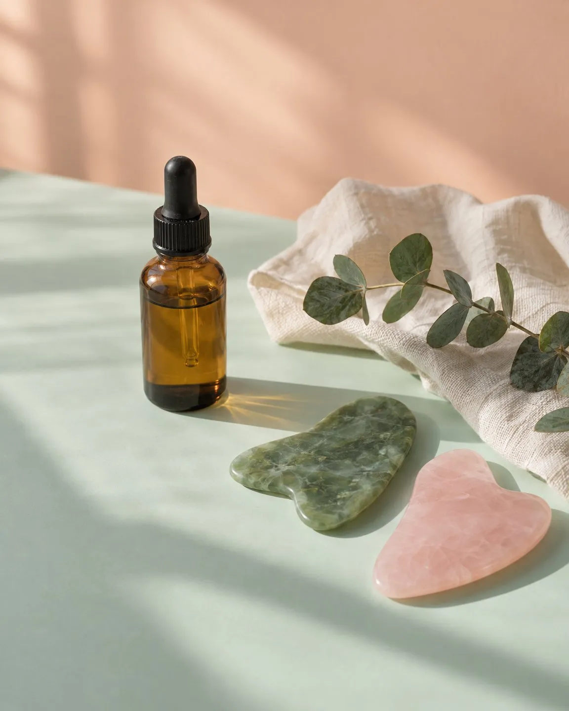Jade and rose quartz gua sha stones, a dropper bottle of facial oil, linen and eucalyptus on a mint surface